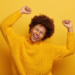 Accomplishment and success concept. Joyful Afro woman raises arms, feels glad and relaxed, tilts head, dressed in casual knitted jumper, laughs from happiness, celebrates victory, isolated on yellow